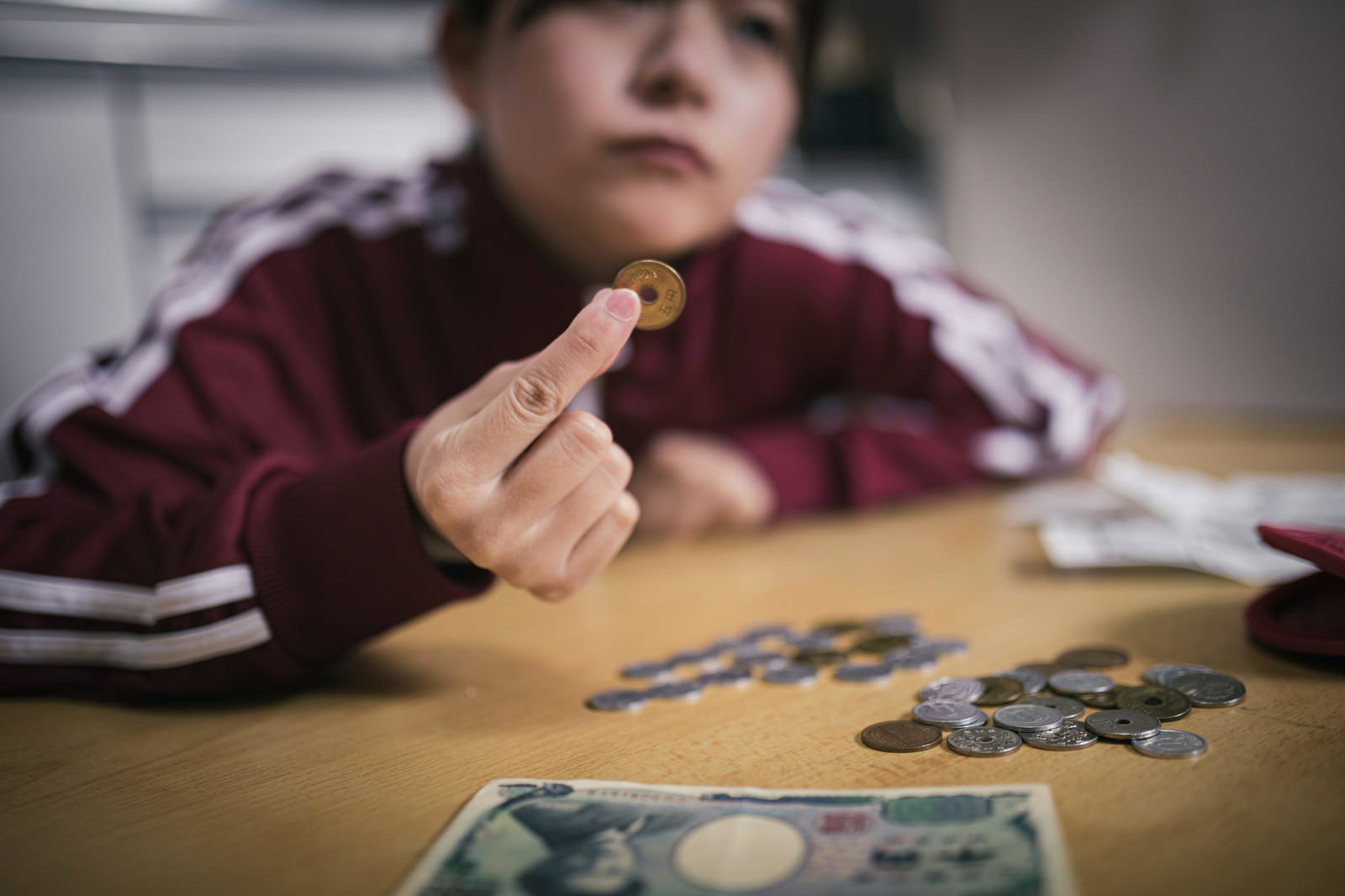 赤いセーターを着た女性が机の上で五円玉を指でつまんで持っている手元［モデル：千歳］
