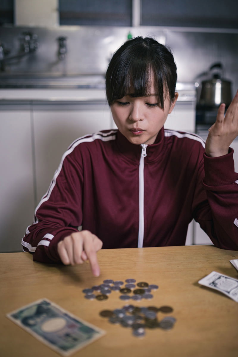 赤いジャージを着た女学生がテーブルで小銭を数えている［モデル：千歳］