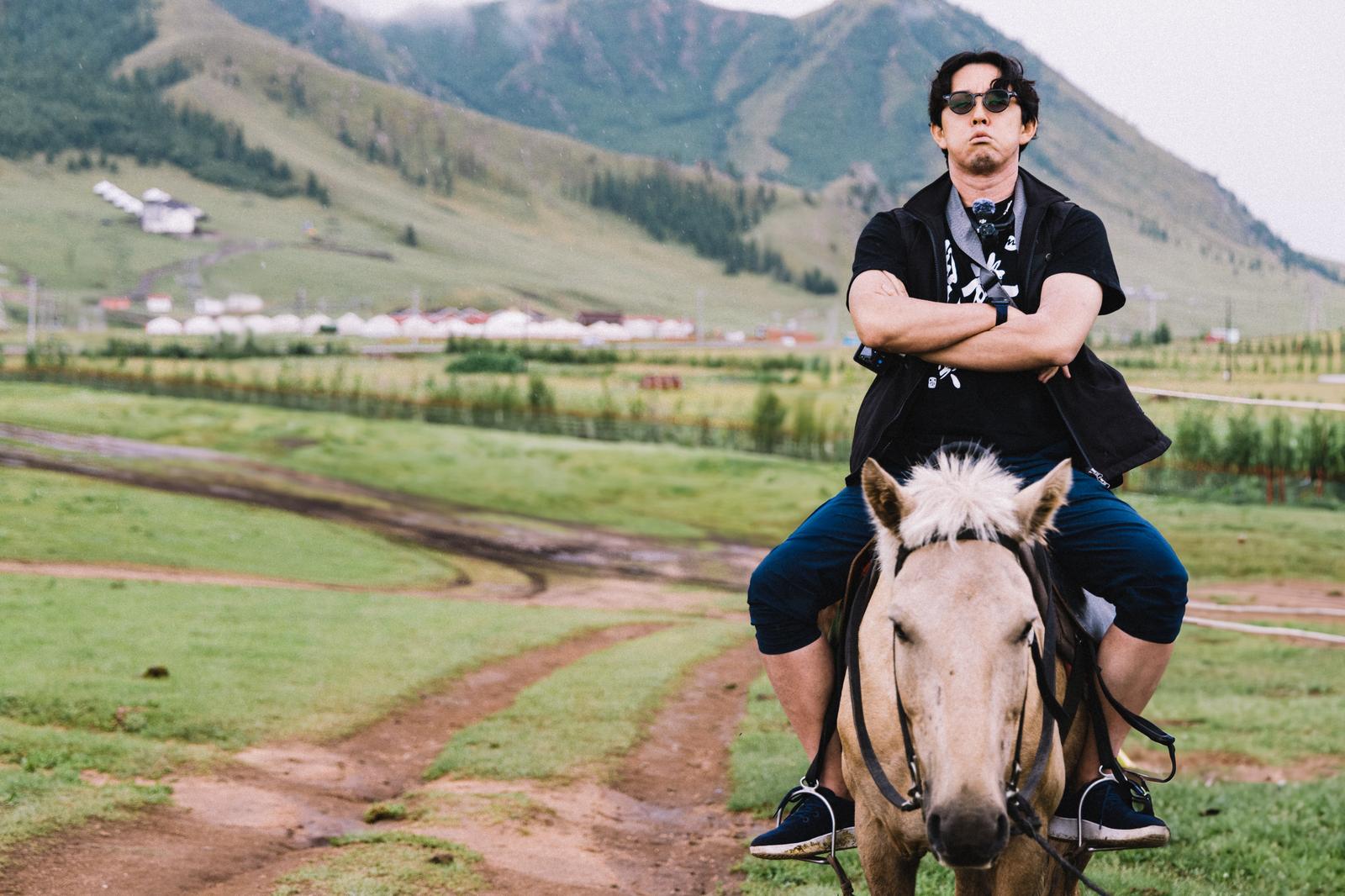 モンゴルの草原で馬に乗り腕を組む男性旅行者、背景に山脈［モデル：もーりー］