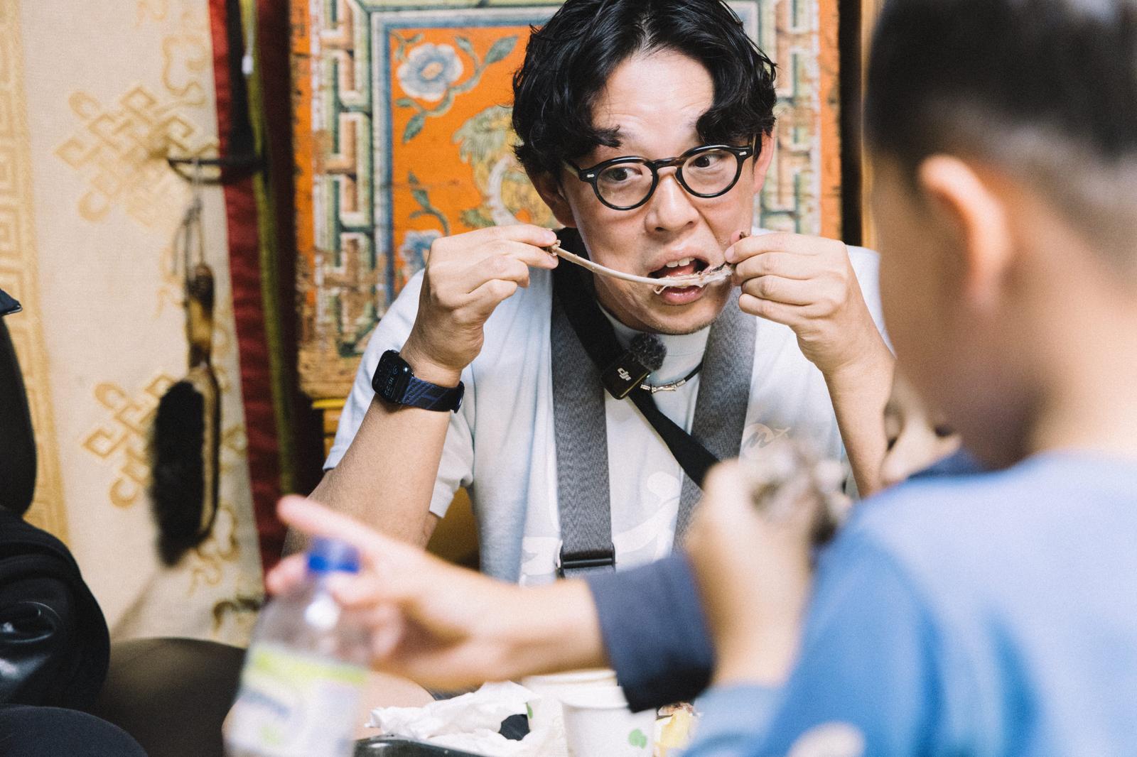 羊肉を食べるYouTuberの男性が箸を持ち、モンゴルの装飾背景で食事中［モデル：もーりー］