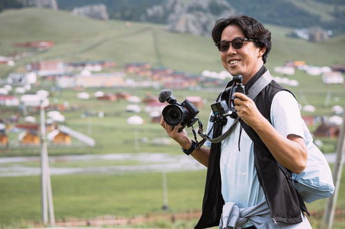 モンゴルの草原に建つ建物を背景に複数の機材を持つ旅行系YouTuber