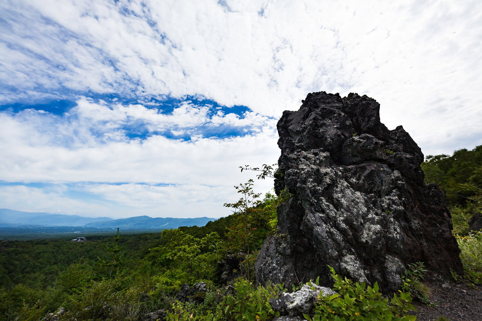 黒い溶岩の岩が樹海に囲まれた鬼押出し園。背景に山々と青空が広がる景観