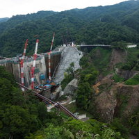 緑の山々に囲まれた建設工事中の八ッ場ダムとクレーンの写真