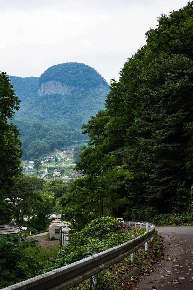 山間部の坂道とガードレール、背景に丸岩の山が見える風景