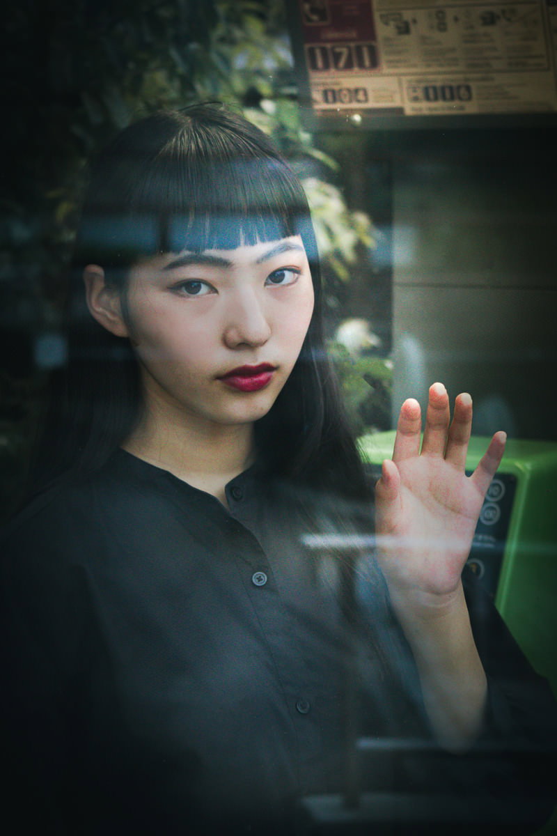 電話ボックスのガラス越しにこちらを凝視する黒髪の女性［モデル：なんこ］
