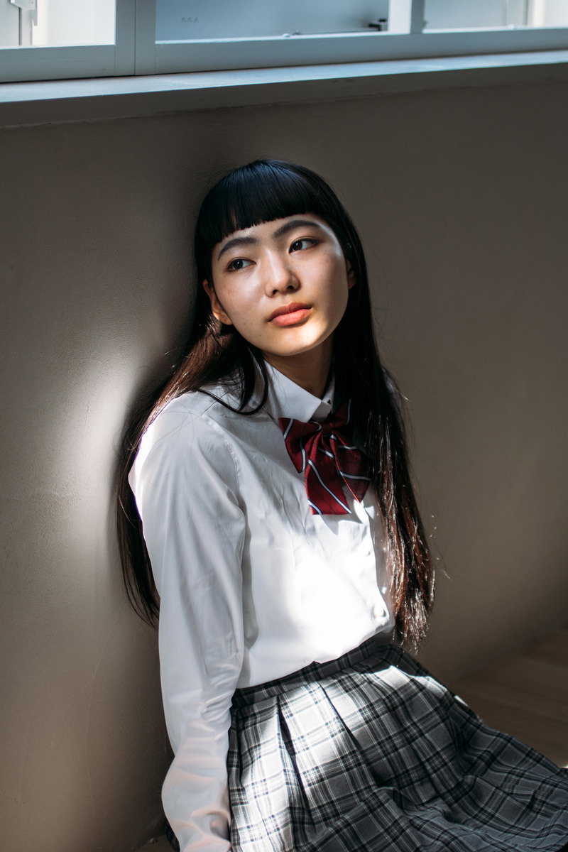 壁にもたれて窓の外を見つめるオン眉の女子高生のポートレート［モデル：なんこ］