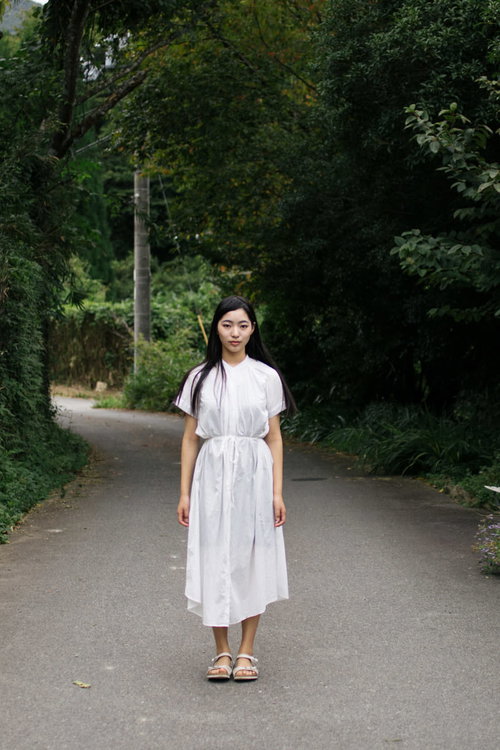 緑に囲まれた道で棒立ちするワンピース姿の女性