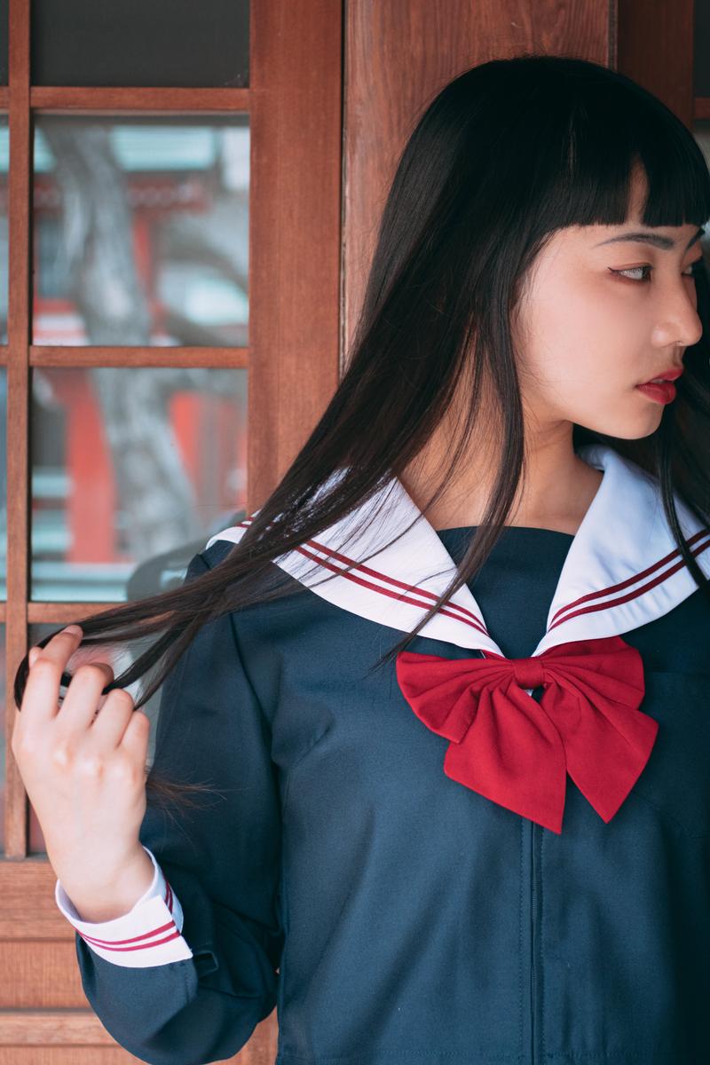 木戸の前で横顔を見せ、長い黒髪に手を添えるセーラー服姿の女子高生［モデル：なんこ］