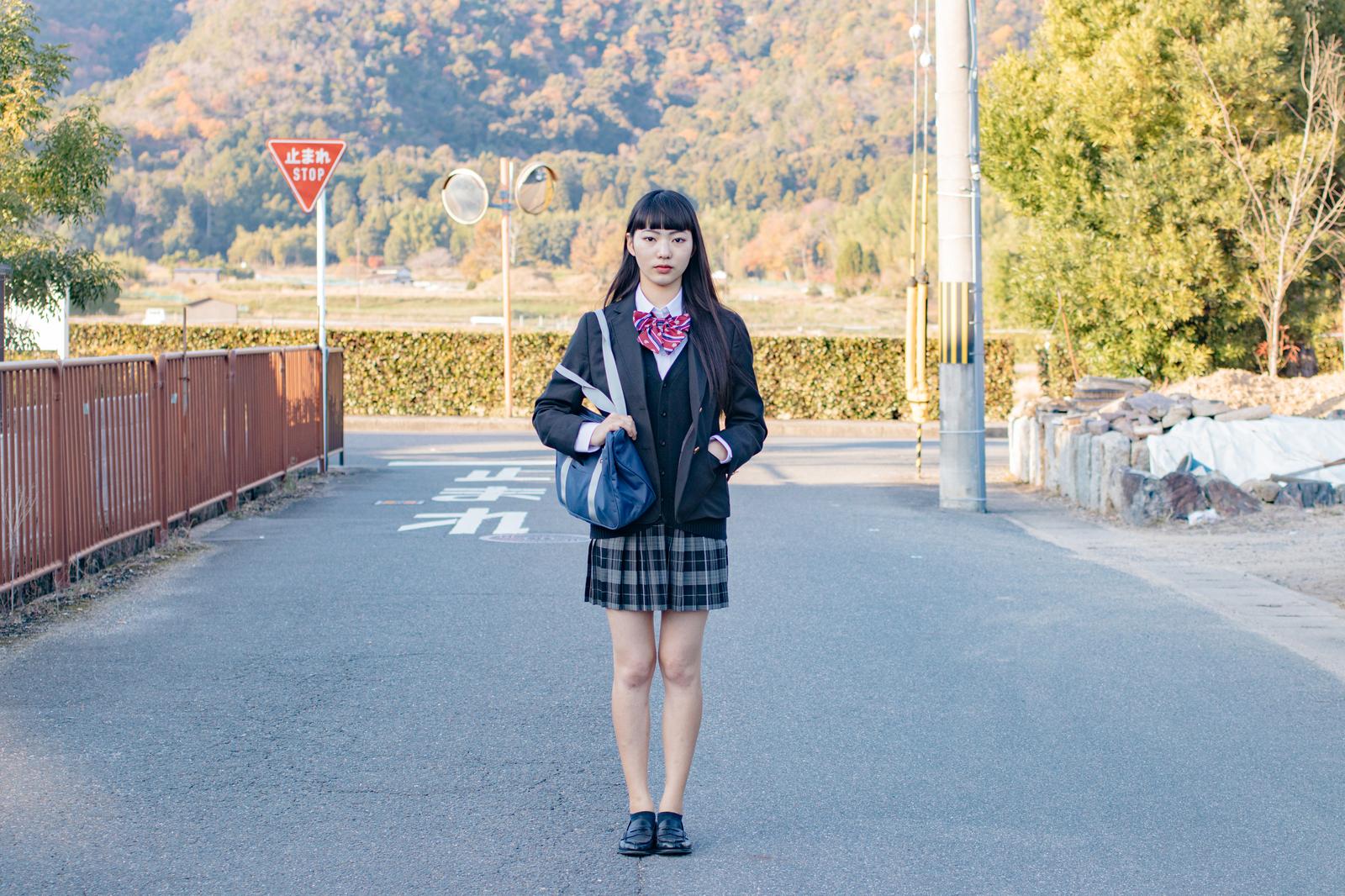 赤いリボンの黒いセーラー服を着た女子高生が山間地の通学路に立っている［モデル：なんこ］