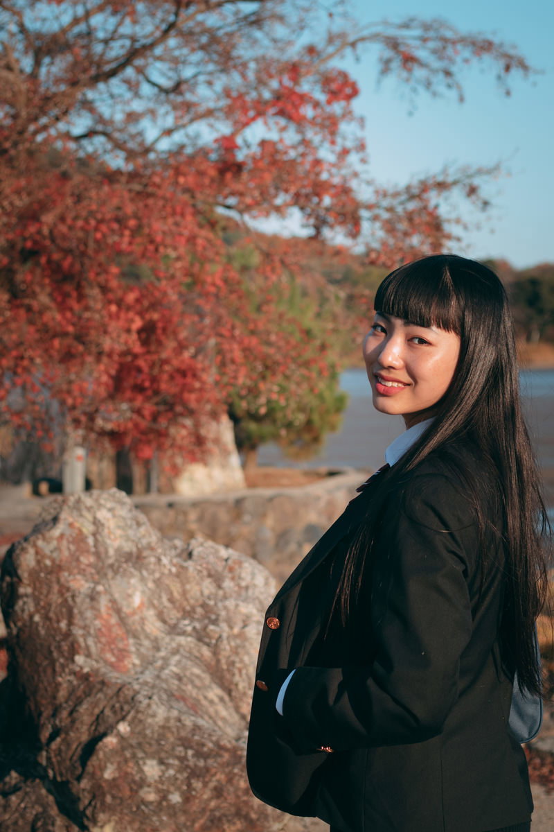 紅葉した木々の前で、黒いコートを着て後ろを振り返る女性の笑顔［モデル：なんこ］