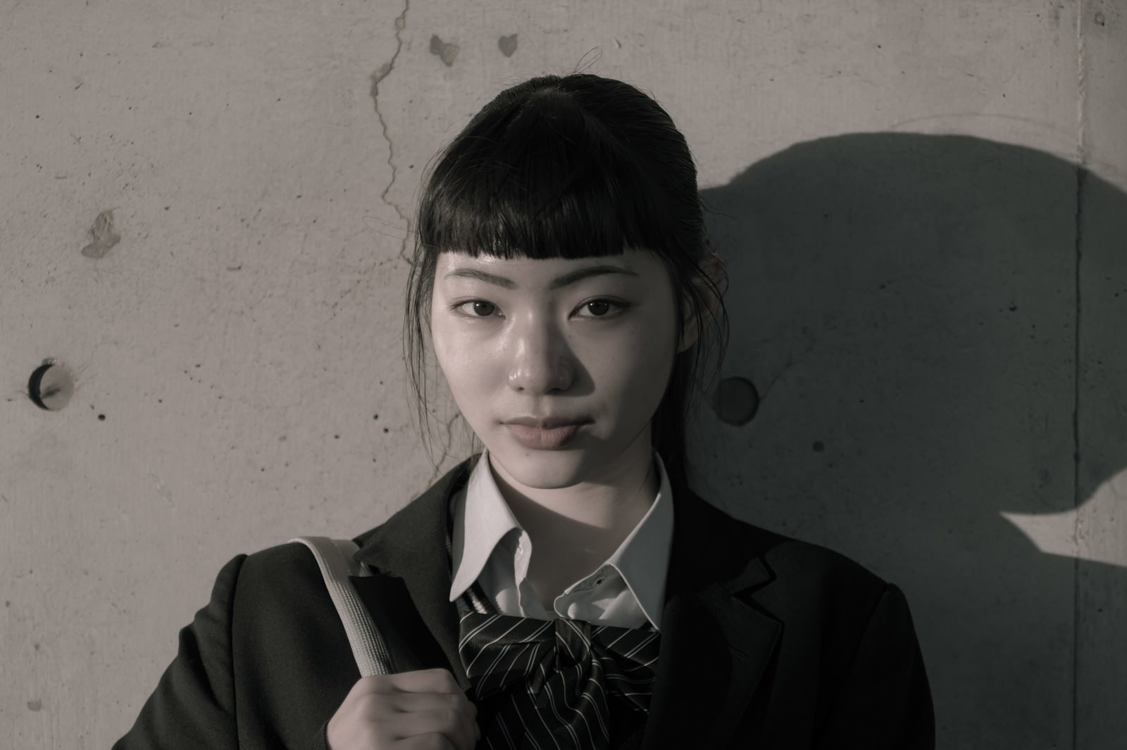 壁を背景に立つ学生服の女子高生と、壁面に映る影［モデル：なんこ］