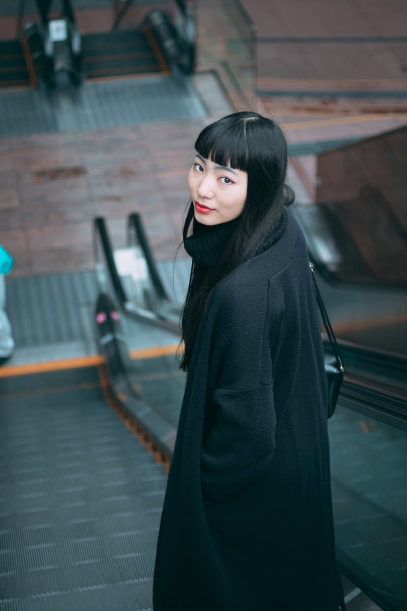 エスカレーターで後ろを振り返る黒いコートを着た女性［モデル：なんこ］