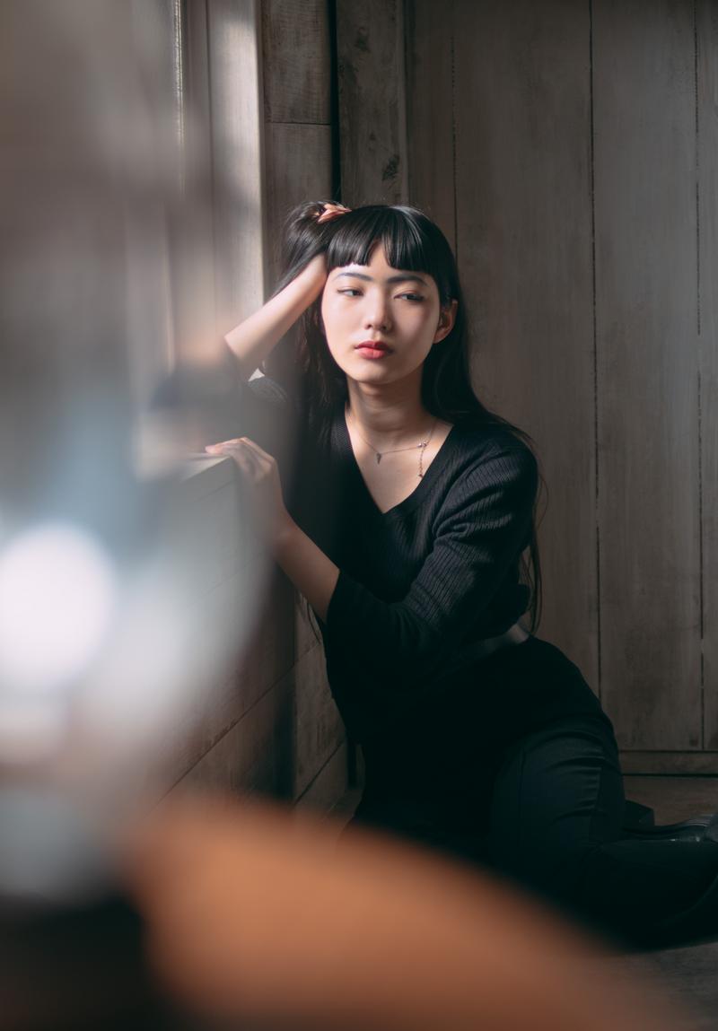 窓辺の自然光の中、木製の壁に寄りかかり思いに耽る黒いセーターの女性［モデル：なんこ］