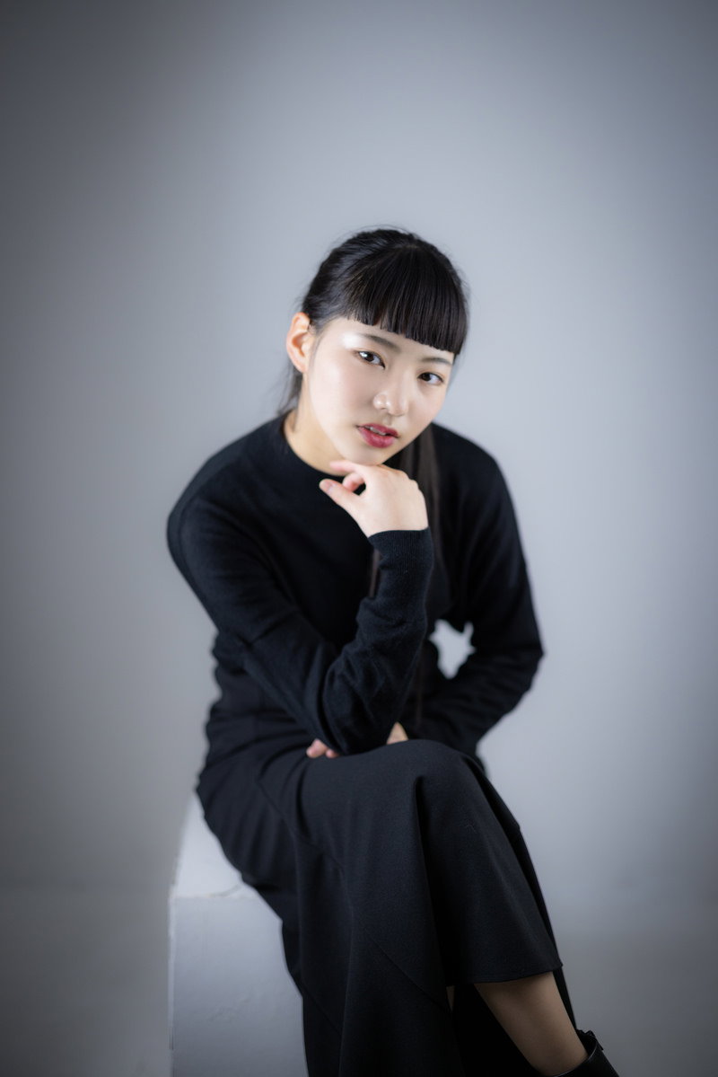 黒いブラウスで足を組んで頬杖をつき澄ました表情の女性［モデル：なんこ］