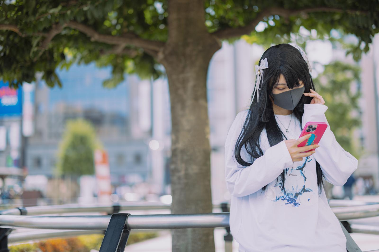 駅前の柵に座ってピンク色のスマートフォンを見る黒いマスク姿の女性［モデル：緋真煉］