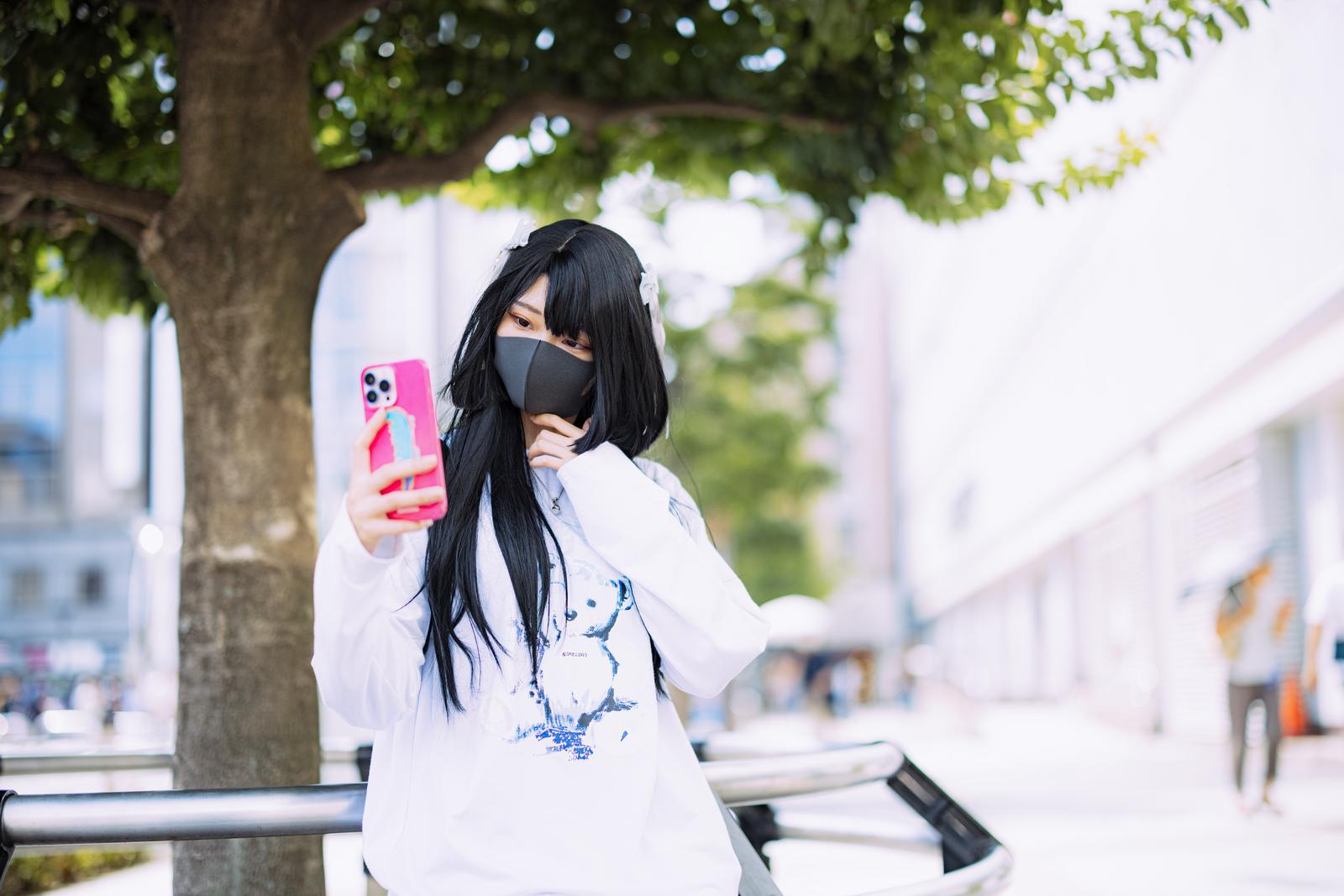 マスク姿でピンクのスマホを持って自撮りする黒髪の女性［モデル：緋真煉］