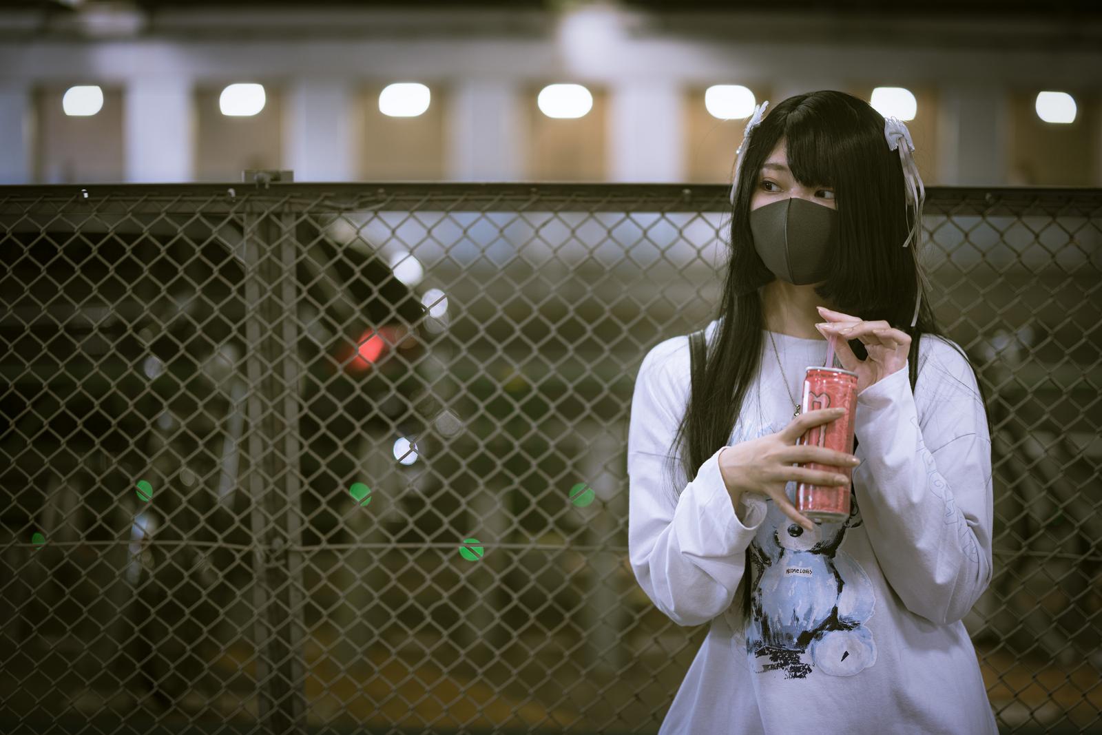 マスクを着けたピンモン女子が夜間の駅のフェンス前でエナドリを持って立っている［モデル：緋真煉］