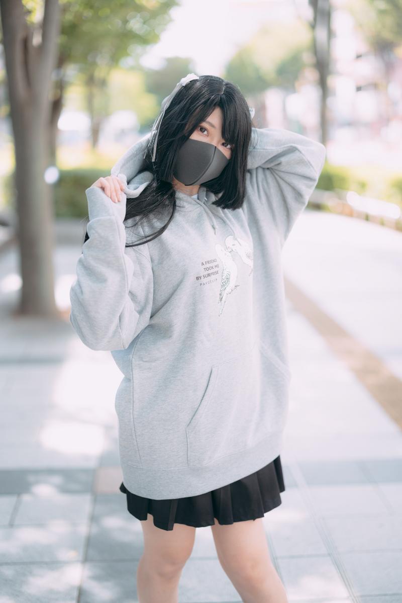 グレーのパーカーを着たマスク姿の女性が樹木の並ぶ道路に立っている［モデル：緋真煉］