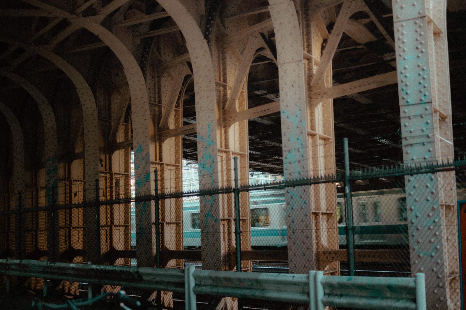 リベット打ちされた鉄骨アーチ構造の高架下と金網フェンス越しの電車