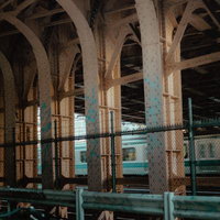 リベットで補強された鉄骨アーチと金網フェンス越しの電車の写真