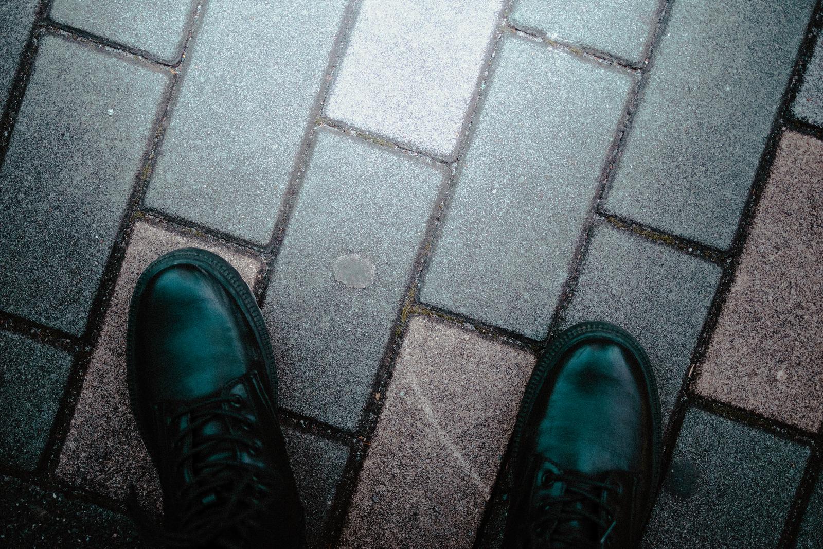 濡れた石畳の歩道に立つ黒い革ブーツの足元を上から撮影した写真