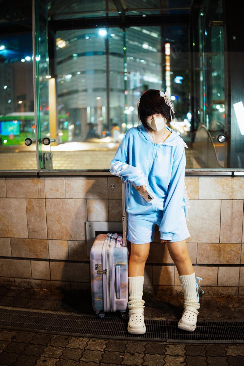 夜景のビル前でキャリーケースを持つ薄紫色のパーカー姿の女性［モデル：緋真煉］