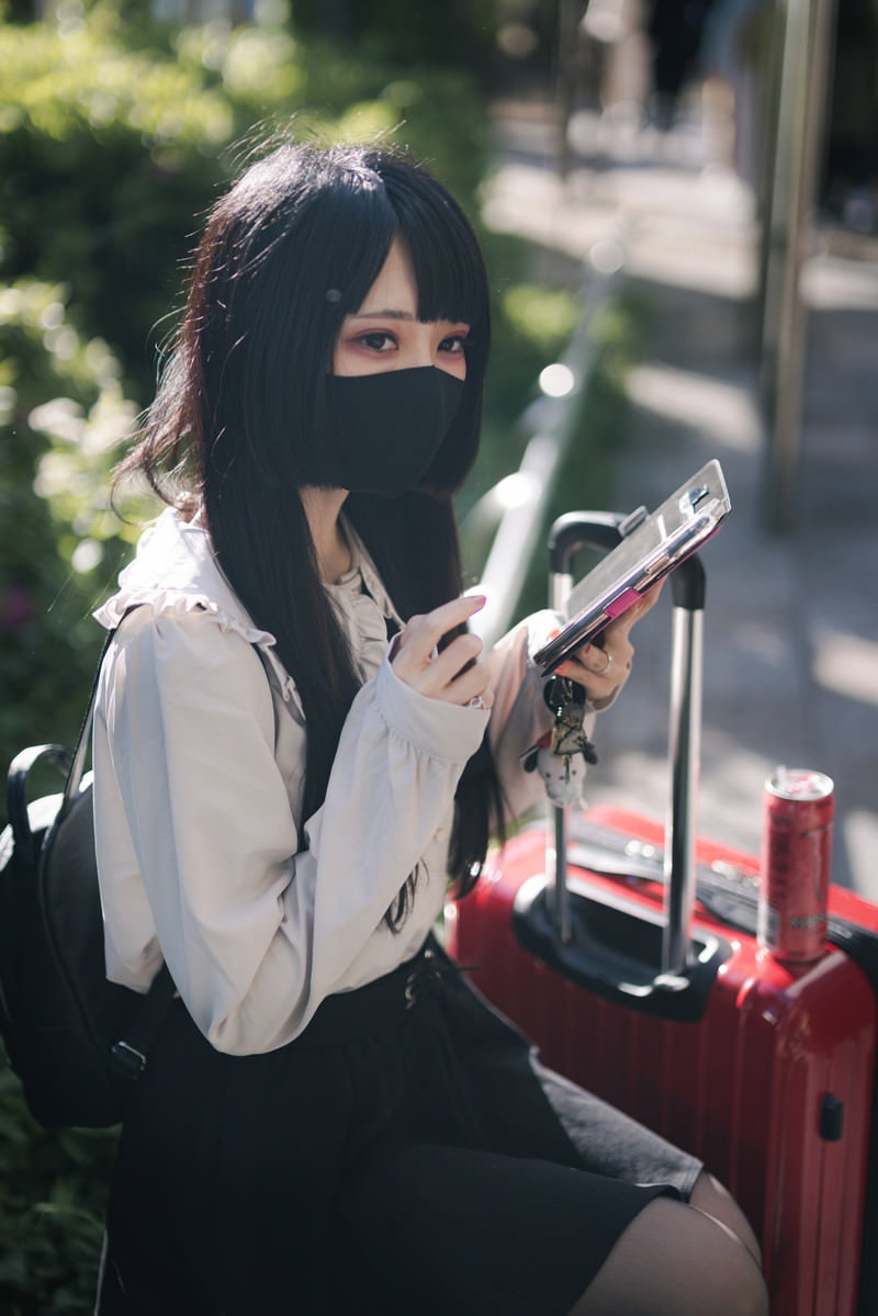 キャリーケースの横に座りスマートフォンを見るマスクをした女性［モデル：緋真煉］
