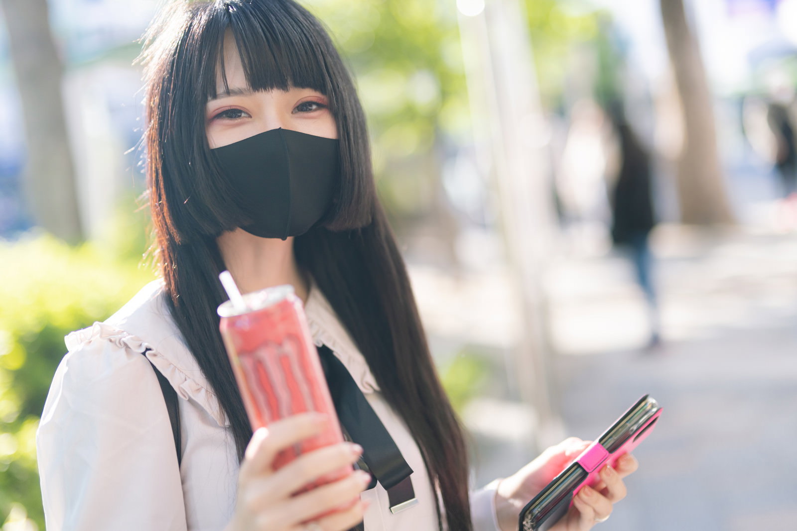 黒マスクをして白い服を着た女性がスマートフォンと飲み物を持っている［モデル：緋真煉］
