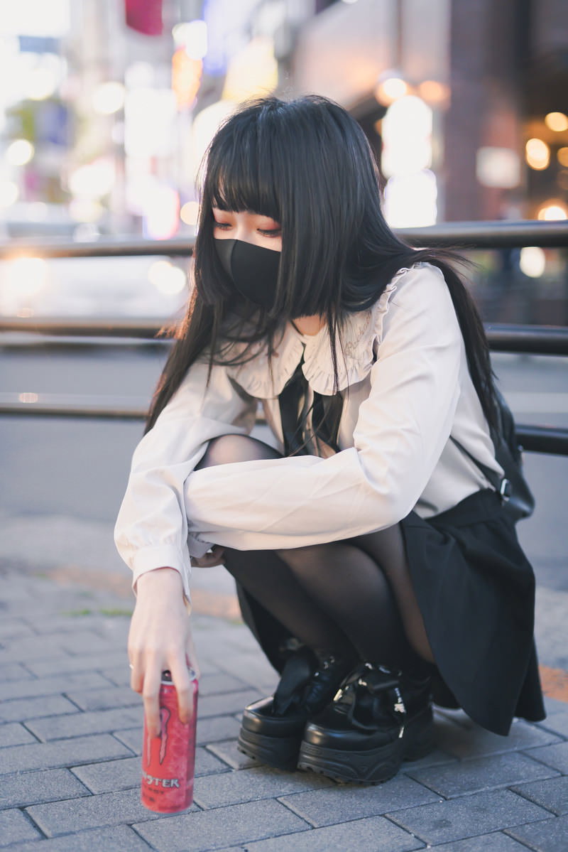 夜の路上でしゃがみこんでいる黒いマスクを着けた女性［モデル：緋真煉］