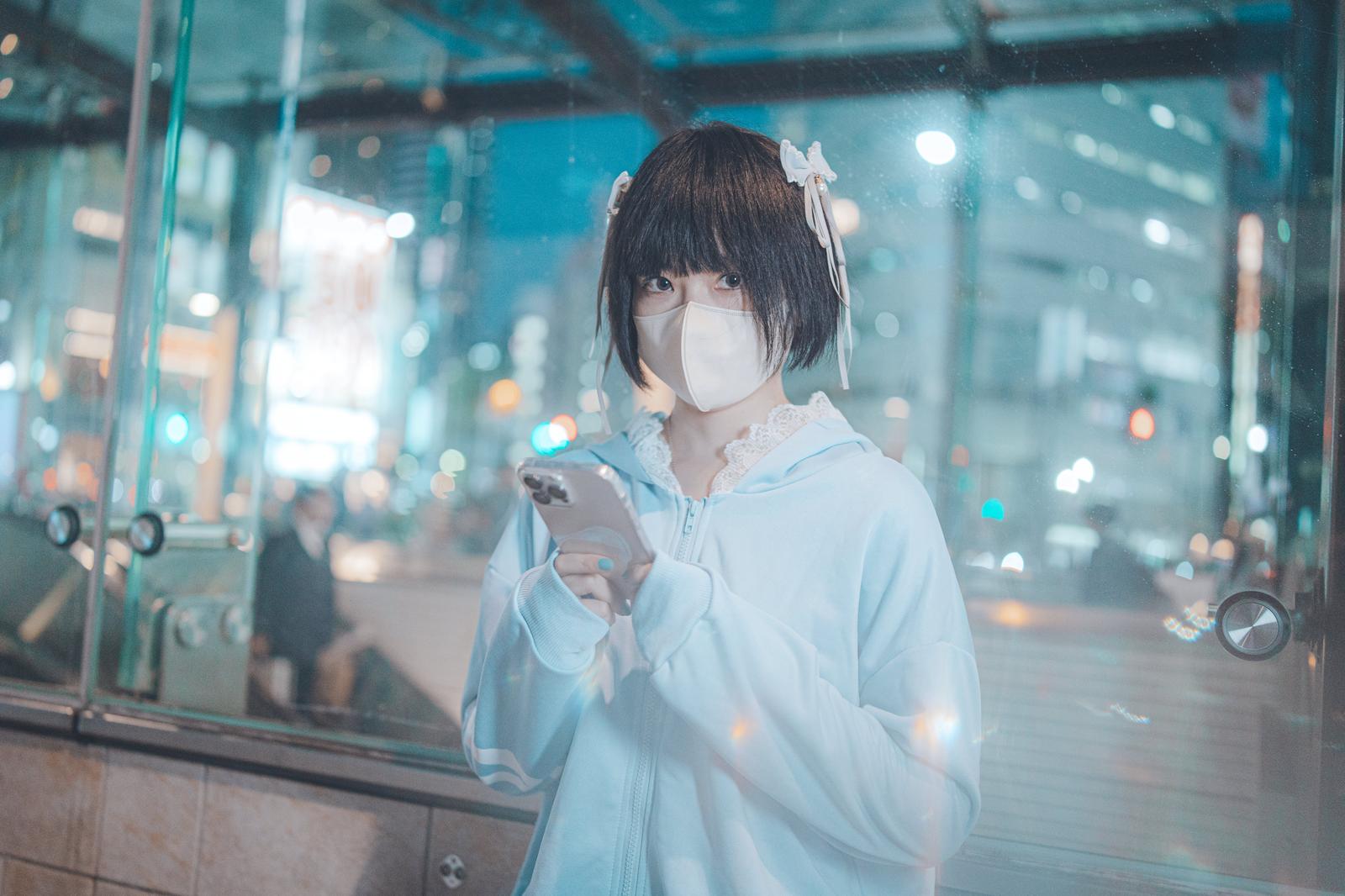 夜間の駅前でマスクをしてスマートフォンを見つめる若い女性［モデル：緋真煉］