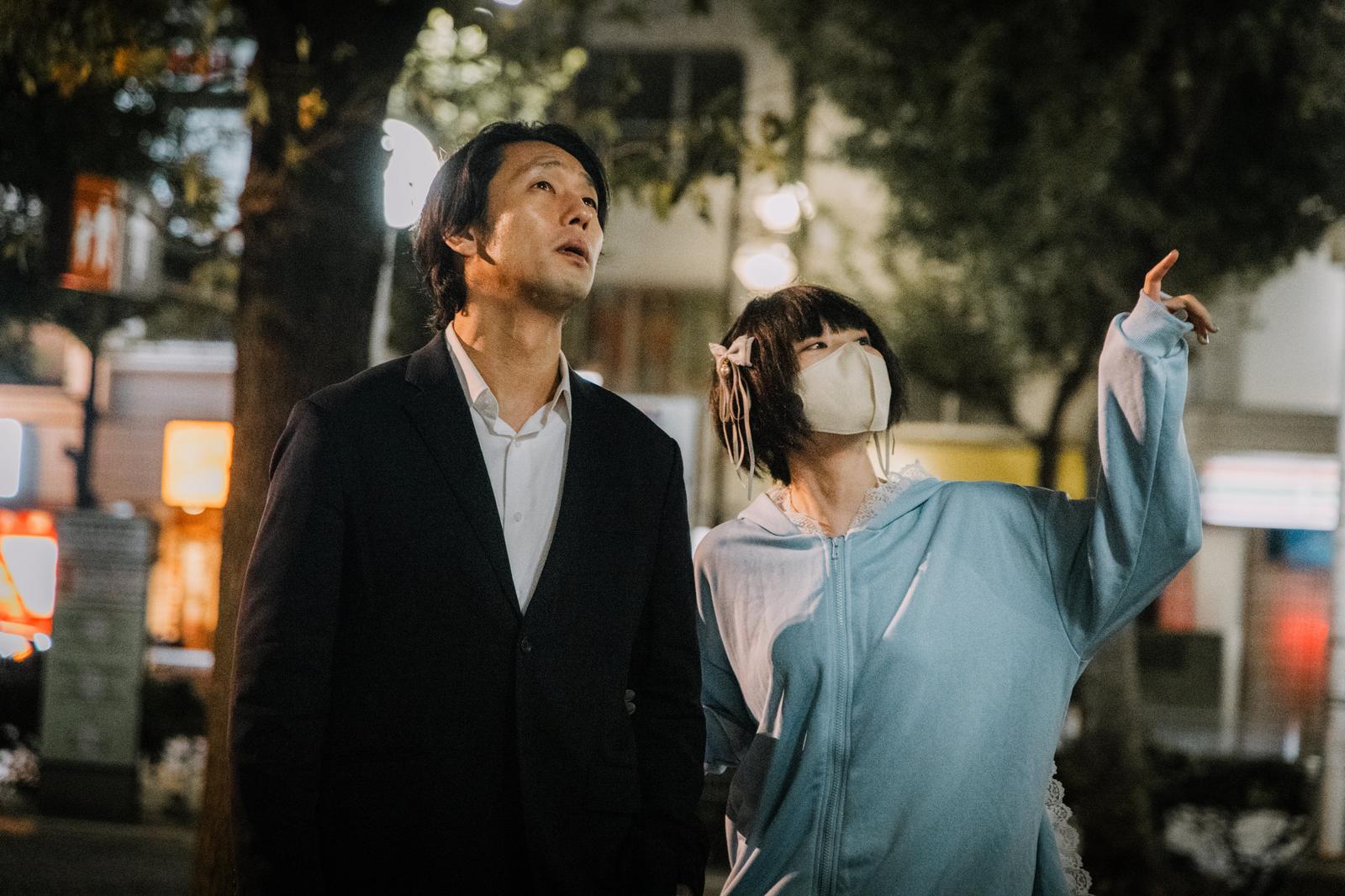 夜間の繁華街でスーツ姿の男性と水色ジャケットのマスク女子が並んで歩く［モデル：大川竜弥 緋真煉］