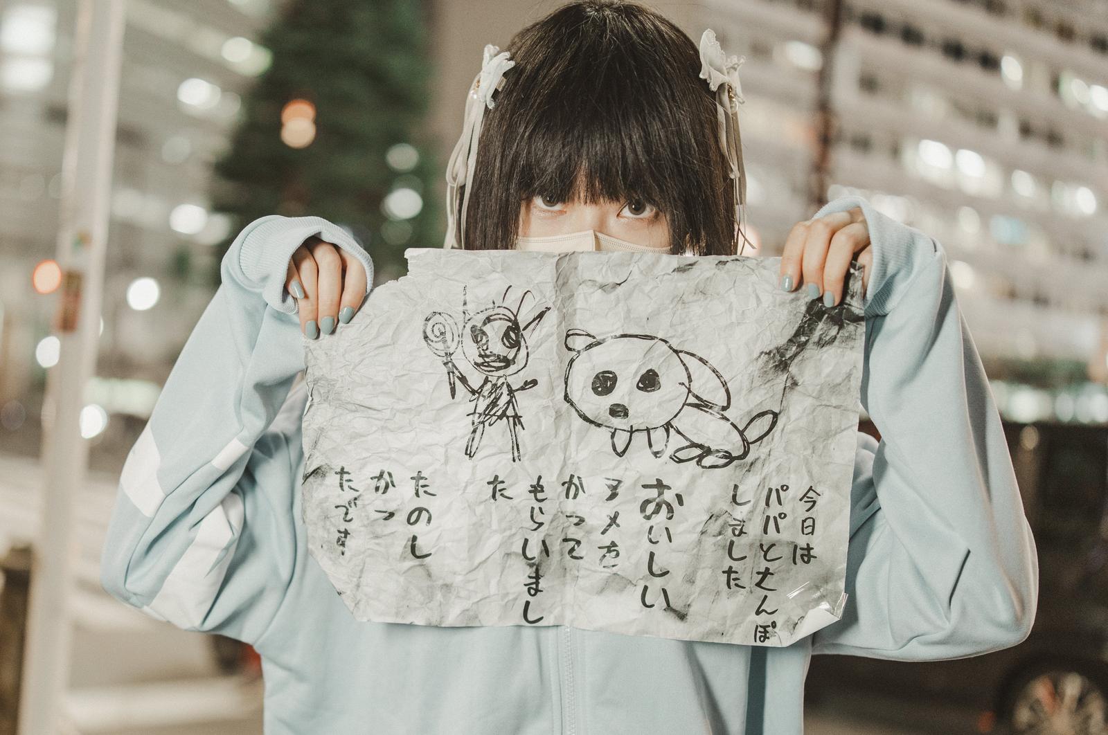 A girl in a blue hoodie is holding up a white sheet of paper with handwritten illustrations and Japanese text in a nighttime city street