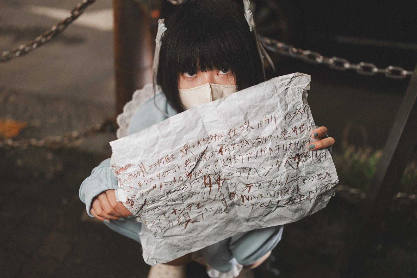 手書きの怪文書を両手で掲げてしゃがむマスク姿の黒髪女性