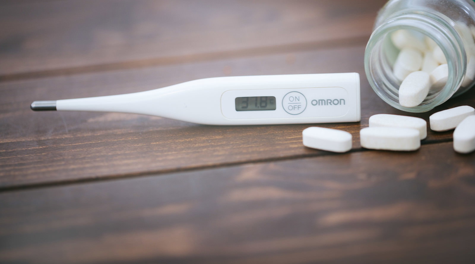 木製テーブルに置かれたomron製デジタル体温計と白い錠剤、背景にグラス