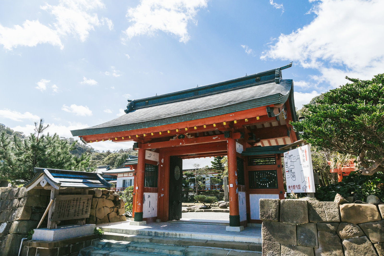 青空の下、鵜戸神宮社務所に続く朱塗りの門と緑豊かな境内