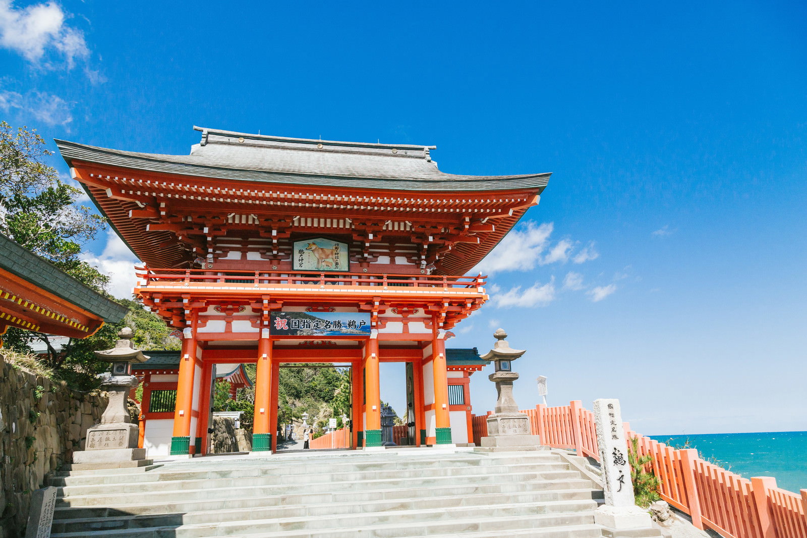 石段の上にそびえる鵜戸神宮の朱色の楼門と青空、右手に広がる海