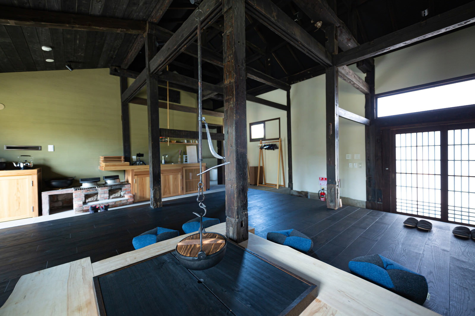 黒い梁が交差する古民家の広い室内に、中央に青い囲炉裏と周囲に青い布団が配置されている