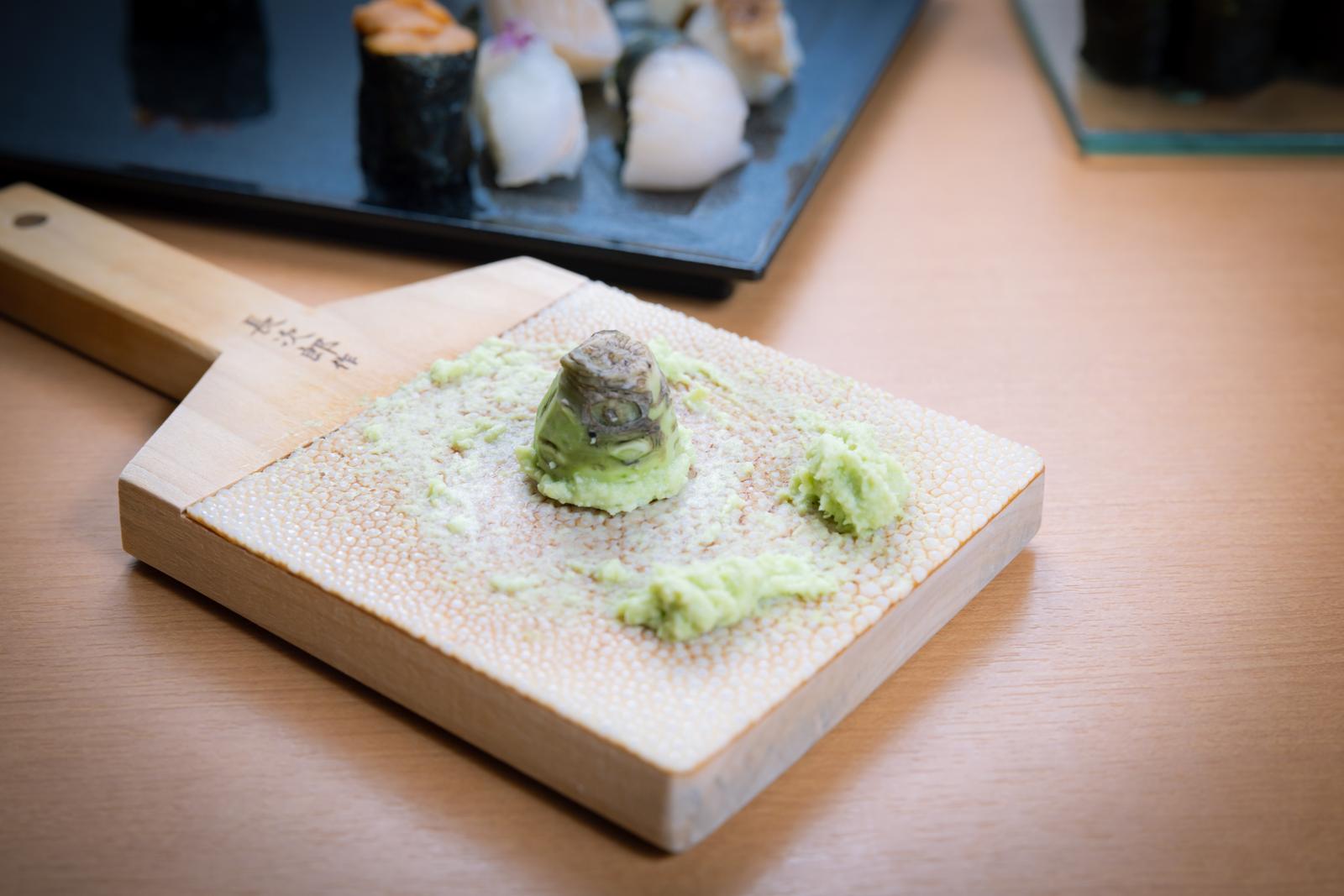 木製のおろし金にすりおろされた本わさび、奥には握り寿司が見える