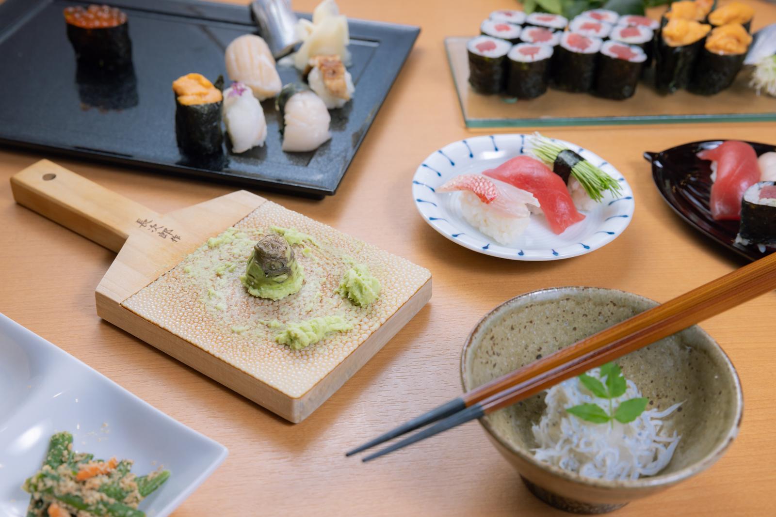 木製のしゃもじに本わさびが乗せられ、複数の皿に寿司と刺身が並ぶ食卓