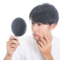 手鏡で頬のニキビを確認するセルフチェック中の男性の写真