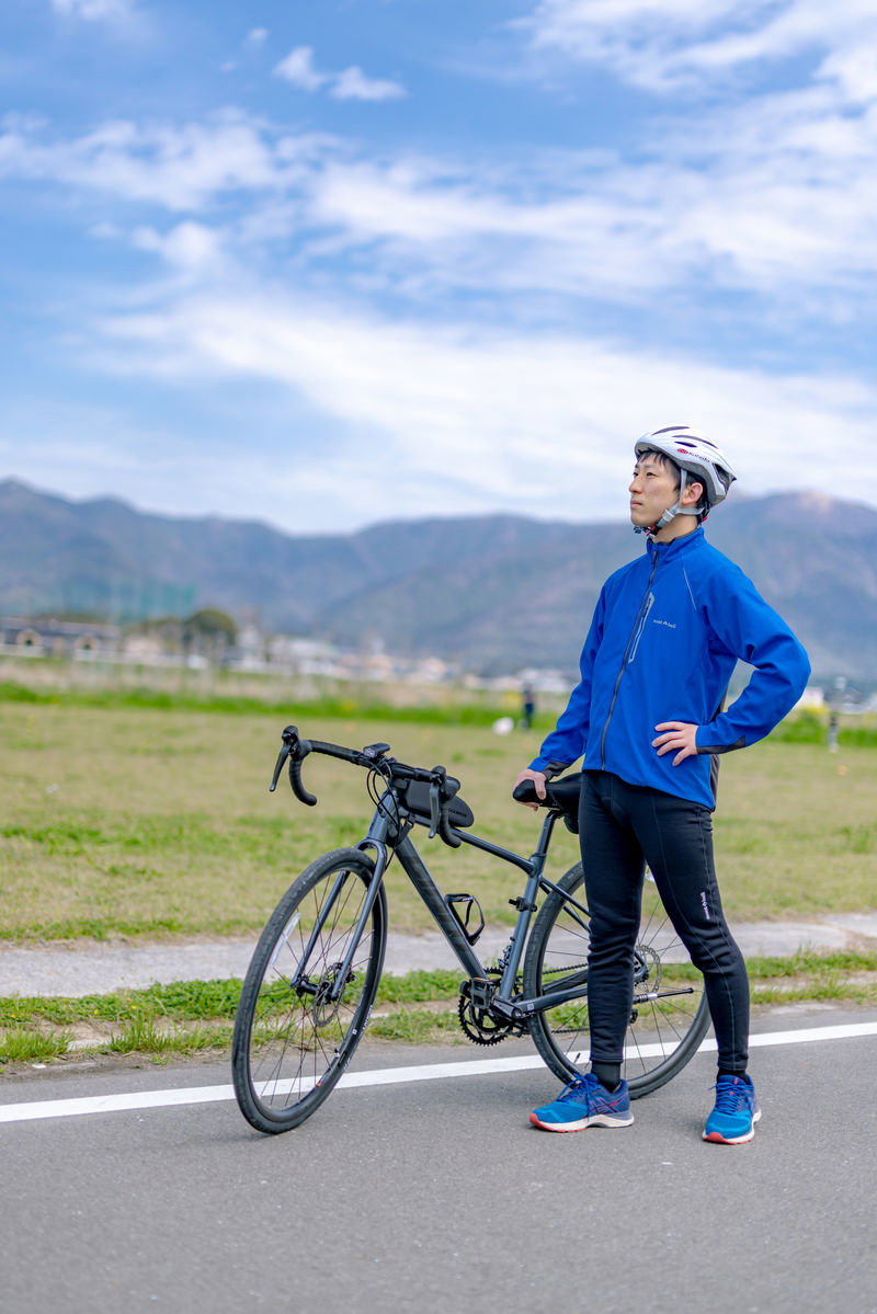 青空と福智山系を背景にロードバイクを持つ青いウェアの男性［モデル：モデルリリース］