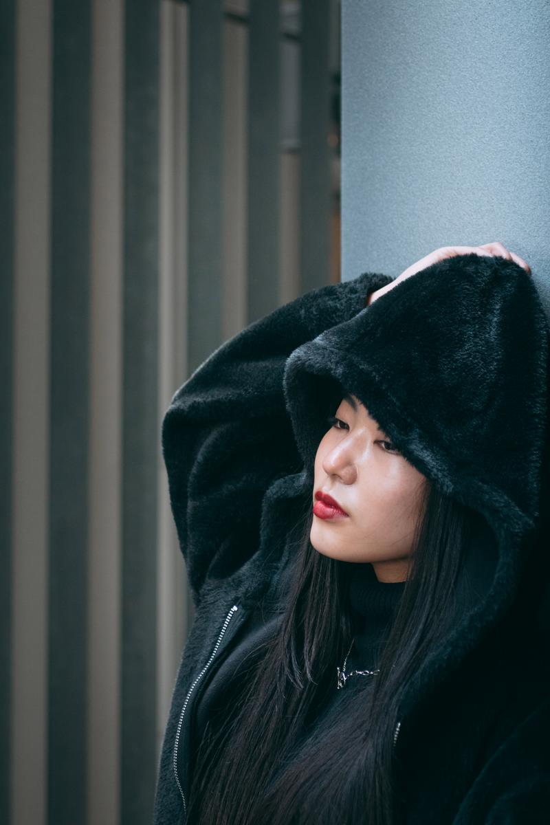 黒いパーカーのフードを被り赤い唇メイクの女性が横向きで立つ［モデル：なんこ］