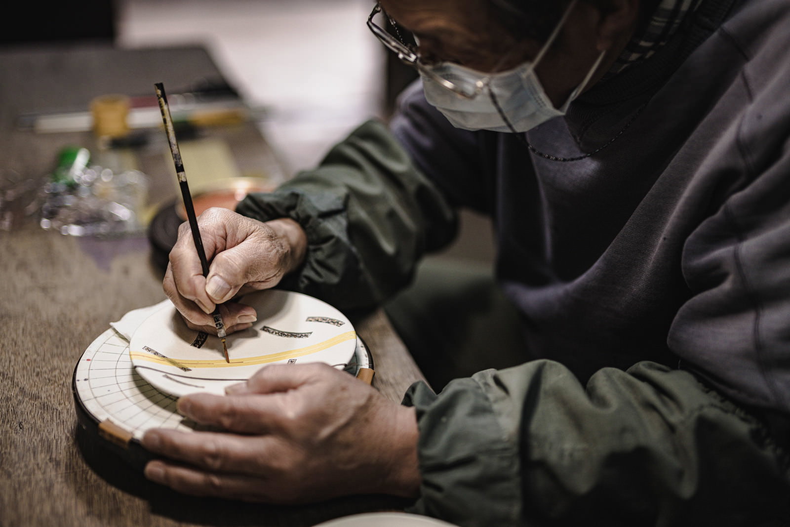 緑の作業着を着た職人が白い陶器を両手で持ち絵付け作業をする手元［モデル：モデルリリース］