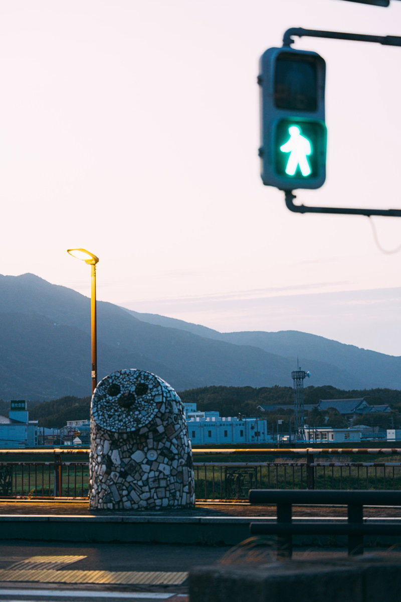 青信号が点灯する横断歩道とふくろう像、夕焼けの山並み