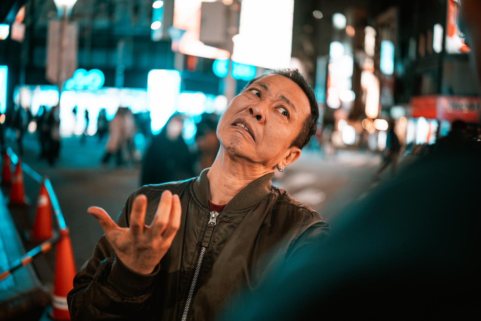 Middle-aged man in a black jacket showing an angry expression with one hand raised in a neon street (Neon gai)