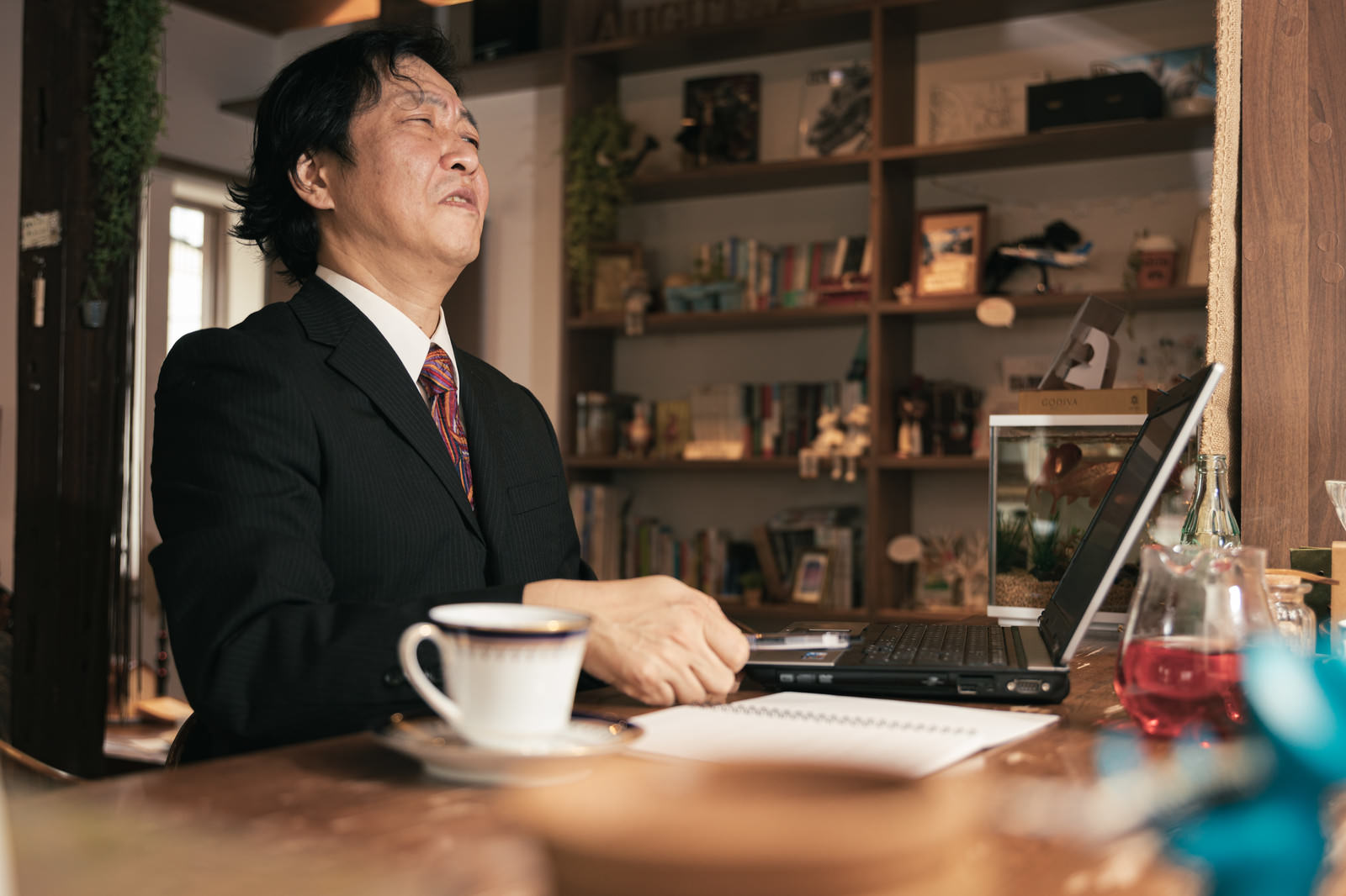 カフェでノートパソコンを見ながら頭を抱えて困った表情を浮かべるスーツ姿の男性［モデル：のせさん］