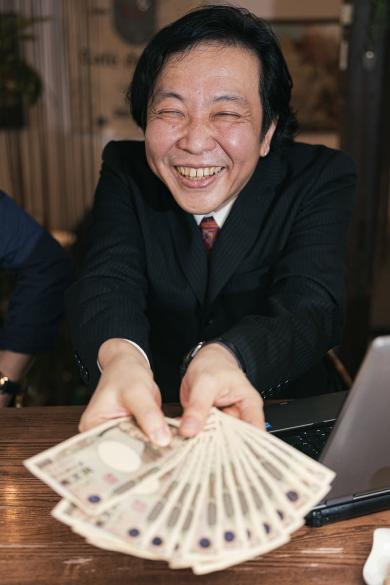 テーブルに万札を広げて満面の笑みを浮かべるスーツ姿の男性［モデル：のせさん］
