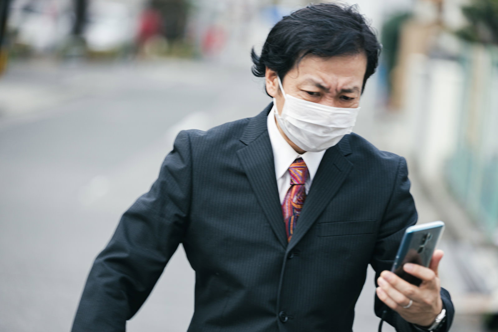 マスクを着用してスマートフォンを見ながら歩く黒いスーツの会社員男性［モデル：のせさん］