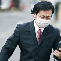 マスクをしながらスマートフォンを見て外出する会社員の写真