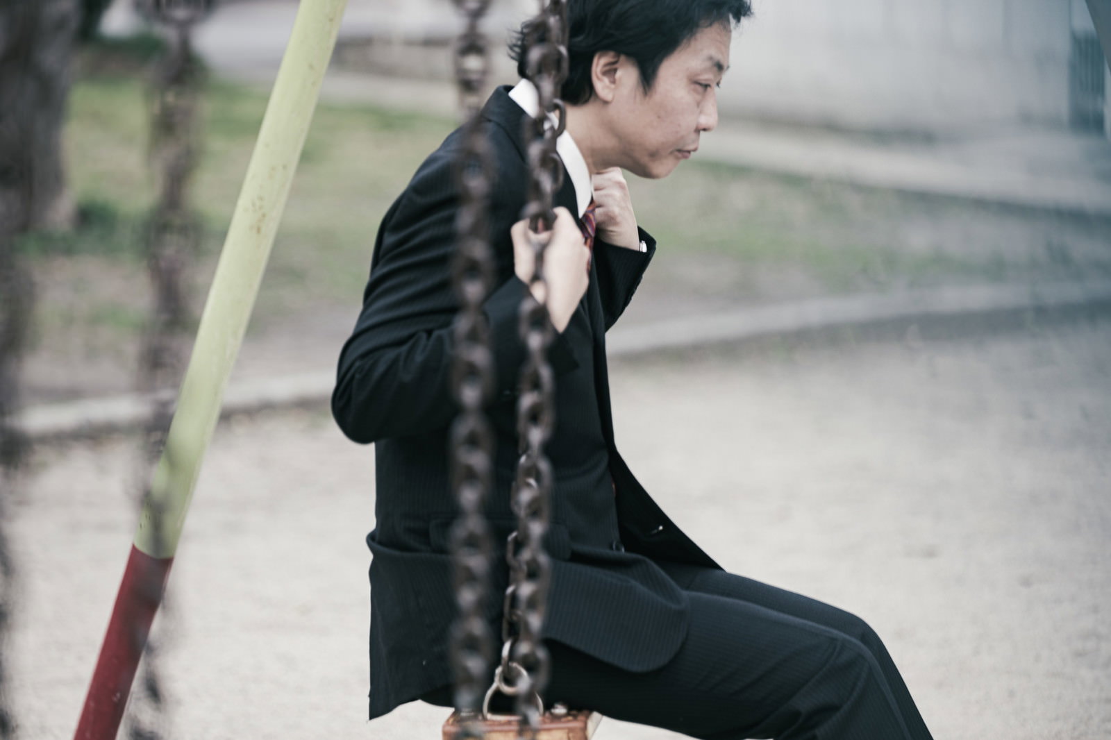 公園のブランコに座り憂鬱そうな表情を浮かべる黒いスーツ姿の男性［モデル：のせさん］
